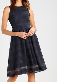 Marinblå ärmlös klänning med ett strukturerat randmönster, figursydd bodice och utsvängd kjol, med längd nedanför knäna och en subtil lyster.