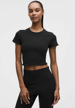 Kvinde med mørk hud og håret sat tilbage bærer en sort ribbet cropped top med flæsekant og sorte leggings med høj talje, stående mod en hvid baggrund.