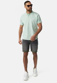 Lys grøn polo-shirt med krave, parret med grå shorts med oprullede manchetter og grønne accenter. Hvide sneakers og solbriller fuldender looket.