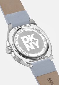 Metalen horlogekast met een zilveren afwerking, grijze achterkant met de tekst "DKNY" en een tweekleurige leren band in lichtblauw en beige. Waterbestendig tot 5 ATM.