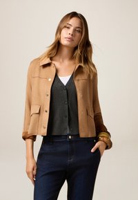 Giacca scamosciata color tan con colletto, bottoni a pressione, due tasche frontali e maniche arrotolate; sovrapposta a un cardigan grigio e jeans blu.