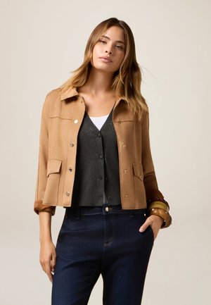 Veste en daim beige avec un col, des boutons-pression, deux poches avant et des manches retroussées ; portée par-dessus un cardigan gris et un jean bleu.