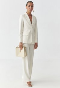 Weißes maßgeschneidertes Blazer und weite Hosen, kombiniert mit einer beigefarbenen Handtasche mit einer Schleifendetail und Riemchensandalen. Glänzende goldene Akzente zieren die Ohrringe.