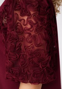 Manche de robe bordeaux avec un tissu texturé agrémenté de rosettes superposées et enroulées sur un fond transparent.