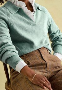Pull vert menthe avec un col en V et des poignets côtelés, superposé à une chemise blanche à col. Pantalon en tartan marron avec une coupe ajustée.