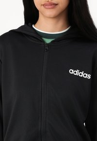 Fekete kapucnis, cipzáros dzseki sima anyagból, bal oldalon fehér "adidas" logóval, alatta pedig egy zöld és fehér réteges pólóval.