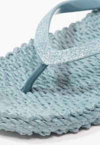 Primer plano de una sandalia tipo chancla azul claro tejida con tiras texturizadas brillantes y una tira entre los dedos.