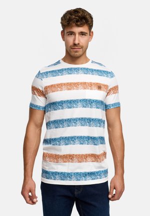 Uomo con maglietta bianca con larghe strisce orizzontali testurizzate blu e arancioni, in piedi contro uno sfondo chiaro uniforme.