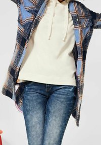 Camisa de franela de cuadros en azul, navy y marrón, superpuesta a una sudadera crema. Unos vaqueros de denim azul completan el atuendo.