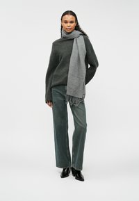 Pelēks adīts džemperis, pelēka šalle ar frizām, zaļi platā kāju bikses, apvienotas ar melnām augstpapēžu potīšu kurpēm. Vienkārša gaismas fona.