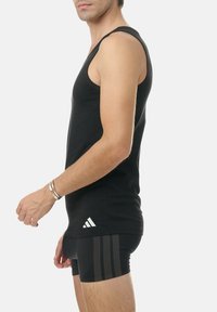 Black sleeveless athletic tank top with a round neckline and Adidas logo on the lower side; model paired with black striped shorts.