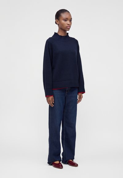 Femme debout portant un pull bleu marine, un jean bleu foncé et des chaussures plates rouges, posant devant un fond gris uni.