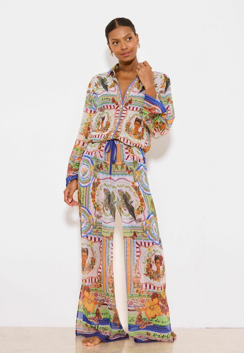 Woman wearing colorful, loose-fit shirt and pants set with artistic portraits, birds, flowers, and text patterns, standing barefoot against plain white background.