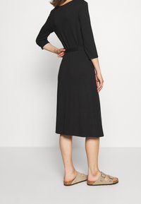 Svart knälång klänning med trekvartsärmar, midjeband och slät textur. Matchas med ljusgrå slip-on sandaler.