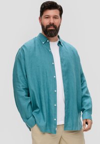 Teal linnen shirt met een knoopsluiting, relaxte pasvorm en lange mouwen over een wit t-shirt. Zachte textuur en minimale hardware-details.