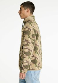 Fleecejack in beige met groen en bruin camouflagepatroon. Voorzien van een hoge kraag, halve rits en zijzakken. Zachte textuur.