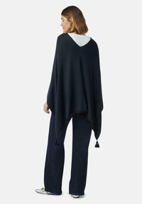 Kobieta w granatowym ponczo z frędzlami, nosząca ciemne szerokie spodnie i srebrne buty, stoi tyłem na tle gładkiej białej ściany.
