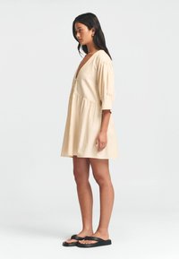 Helles beige Kurzes Kleid mit tiefem V-Ausschnitt, Puffärmel und gestuftem Saum. Getragen mit schwarzen Flip-Flops vor einem schlichten Hintergrund.