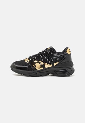 Sneaker atletica nera e oro con dettagli in mesh e vernice, lacci riflettenti e suola spessa, con etichette "Versace Jeans Couture".