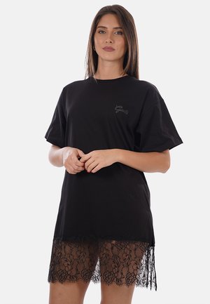 Mujer con el pelo castaño liso lleva un vestido tipo camiseta negro y de gran tamaño con dobladillo de encaje, de pie frente a un fondo claro y liso.