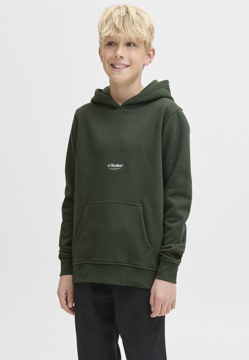 Garçon blond souriant portant un sweat à capuche vert foncé avec poche avant et pantalon noir, debout devant un fond clair uni.