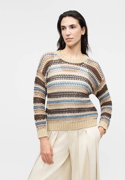 Γυναίκα με ανοιχτόπλεκτο πουλόβερ με ρίγες σε μπεζ, καφέ και μπλε, και εκρού παντελόνι με φαρδιά μπατζάκια, στέκεται με το ένα χέρι στην τσέπη.