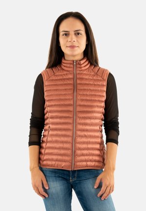 Donna con lunghi capelli scuri che indossa un gilet trapuntato color ruggine sopra un top a maniche lunghe nero e jeans blu, in piedi su uno sfondo bianco.