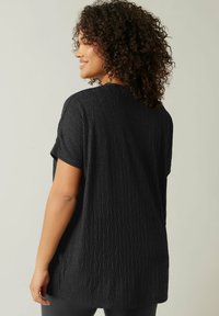 Schwarzes Kurzarmoberteil aus strukturiertem, zerknittertem Stoff mit geometrischem Muster. Lockere Passform mit Rundhalsausschnitt und seitlichen Schlitzen.