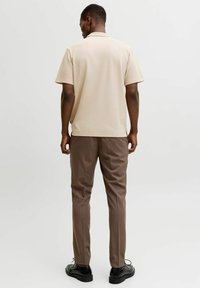 Homme portant un polo à manches courtes beige, un pantalon marron et des chaussures habillées noires, debout de dos sur un fond clair uni.
