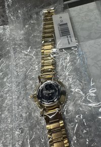 Gold-tone stainless steel watch with a linked band. Features a round black face with silver markings. Packaged in bubble wrap.