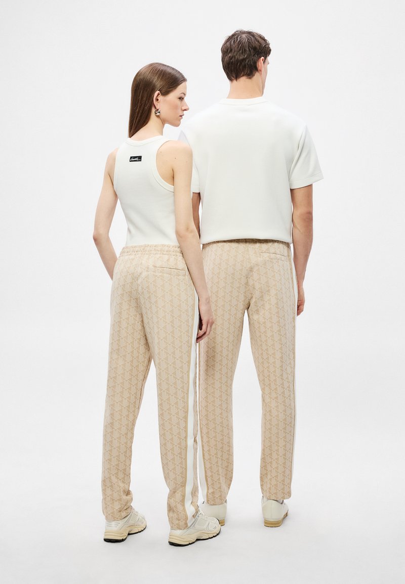 Pantaloni jogger beige con motivo a righe e una striscia bianca, abbinati a un top aderente bianco e a una camicia a maniche corte, visti da dietro.