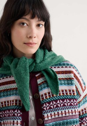 Woman with dark hair wearing a colorful patterned cardigan and green sweater tied around her shoulders on a white background.
