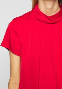 Robe rouge vif avec un col bénitier, des manches courtes et de légers plis à l'avant, réalisée dans un tissu lisse et léger.