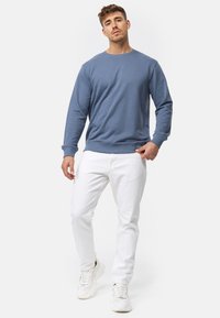 Sudadera de cuello redondo azul con puños y dobladillo de canalé, combinada con jeans blancos y zapatillas. Detalle de logotipo simple en el pecho.
