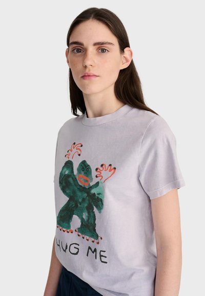 Jeune femme aux longs cheveux bruns portant un t-shirt violet clair avec un monstre vert et les mots "HUG ME" sur fond blanc.