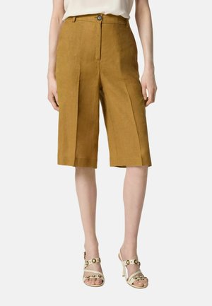 Donna che indossa pantaloncini sartoriali giallo senape fino al ginocchio e sandali beige con tacco alto e borchie dorate, in piedi su sfondo bianco.