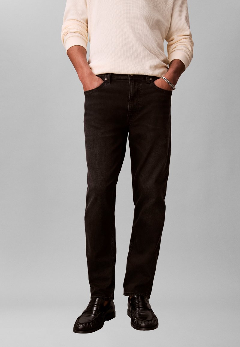 Homme portant un haut beige à manches longues, jean noir, mocassins en cuir noir, mains dans les poches, et bracelet en argent au poignet droit.