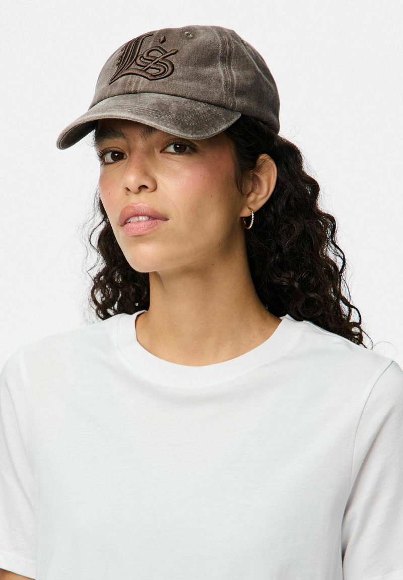 Cappellino da baseball in cotone grigio con logo ricamato, visiera curvata e una texture usurata. Indossato con una maglietta bianca semplice.