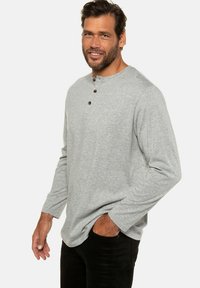Graues Langarm-Henley-Shirt aus weichem Material, mit einer Drei-Knopf-Leiste und dezentem Logo, getragen über schwarzen Hosen.