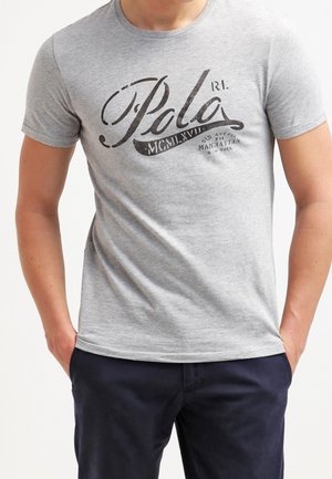 Mand iført en lysegrå Polo Ralph Lauren t-shirt med sort logo tekst og mørkeblå bukser, hænderne i lommerne, stående mod en ensfarvet baggrund.
