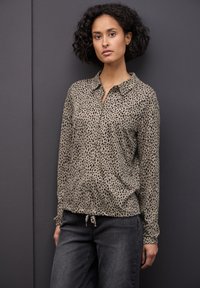 Blusa a maniche lunghe con bottoni di colore beige, con un motivo a macchie nere, realizzata in tessuto morbido, dotata di colletto e fondo con cordino.