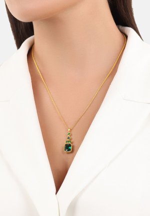 Collana d'oro con un pendente rettangolare di pietra verde, accentuato da piccole pietre verdi disposte in verticale, indossata sopra una camicia bianca.