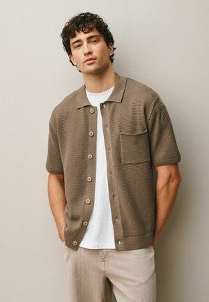 Mann mit lockigem dunklem Haar, der eine braune kurzärmelige Strickjacke mit Knöpfen über einem weißen T-Shirt und beigen Hosen trägt und vor einer schlichten Wand steht.