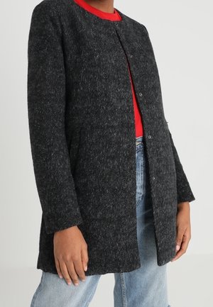 Person trägt einen dunkelgrauen, strukturierten Mantel über einem roten Oberteil und hellblauen Jeans, mit entspannten Händen an den Seiten.