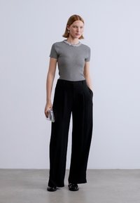 Γκρι ριμπ κοντομάνικο τοπ με κολάρο από χάντρες, συνδυασμένο με μαύρο παντελόνι φαρδιάς γραμμής. Το μοντέλο κρατά μια μικρή ασημένια τσάντα φάκελο.