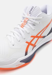 Witte sportieve schoen met oranje en zwarte accenten. Beschikt over een ademende bovenkant van mesh, textuurmateriaal, ingewikkelde veters en een gevoerde zool.