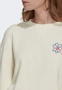Ljuskrämfärgad sweatshirt med avslappnad passform, prydd med en blå och rosa blommönstring på bröstet. Slät textur och synliga sömdetaljer.