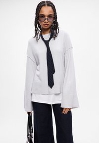 Ανοιχτό γκρι υπερμεγέθες πουλόβερ με φαρδιές μανίκες, συνδυασμένο με λευκό πουκάμισο, συμπληρωμένο με μια ναυτική ριγέ γραβάτα και σκούρο τζιν παντελόνι.