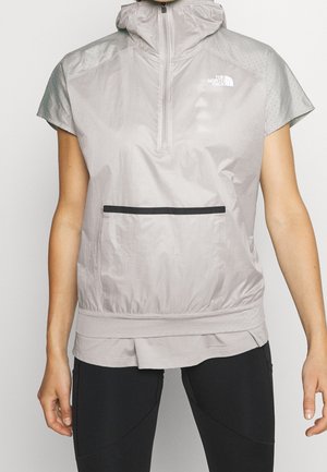 Veste sans manches gris clair à capuche, avec demi-fermeture éclair avant et poche zippée horizontale noire, portée sur un ourlet superposé.