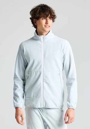 Un homme porte une veste bleu clair zippée à col montant, assortie à un pantalon, sur un fond uni et clair.